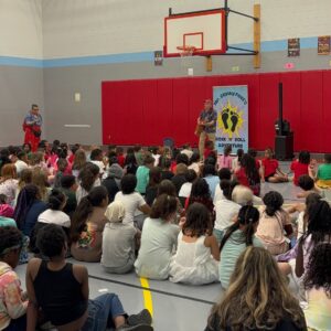 Students sitting in a gym and listening to an artist play music on a guitar